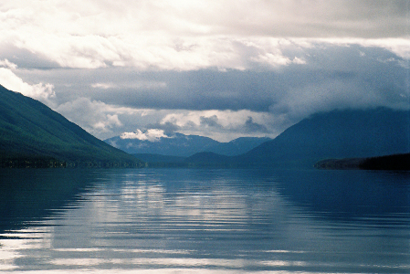 [The entire image is mostly  shades of blue, white, and grey. The wide wake of water from the boat created ripples in the water leading toward a low section between two mountains on either side of the far end of the image. In the very far distance are mountains crossing this low section. The huge amounts of clouds overhead are blocking much of the sunlight.]